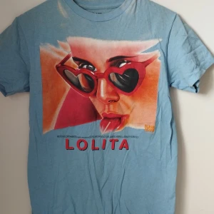 Blå Lolita t-shirt stl S - Snygg ljusblå t-shirt med Lolita-tryck framtill i rött och orange. Tillverkad i 100% bomull för en skön och mjuk känsla. Perfekt för dig som gillar coola filmreferenser och retrostil. Ursäkta att den inte är strykt, håller på att rensa ur garderoben och har inte tid för att stryka.