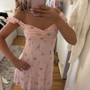 Söt blommig rosa kortklänning - Superfin ljusrosa kortklänning med små blommor i olika färger. Klänningen har offshoulder-ärmar som kan bäras både på och av axeln, rynkad byst och lätt utsvängd kjol. Perfekt för sommarens alla tillfällen!