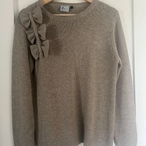 Beige stickad tröja med rosetter  - Vacker beige stickad tröja med rosetter längs axeln!!Tröjan har rund halsringning och långa ärmar. Storlek M men passar även S då den är ganska strechig. Använd 1 gång och fint skick💖