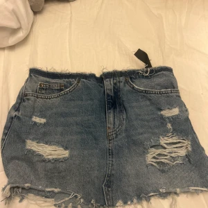 Blå jeanskjol med slitningar från ZARA - Säljer en blå jeanskjol från ZARA i storlek XS. Kjolen har råa kanter och flera slitningar både fram och bak för en trendig, avslappnad look. Klassisk femficksmodell med dragkedja och knapp framtill.