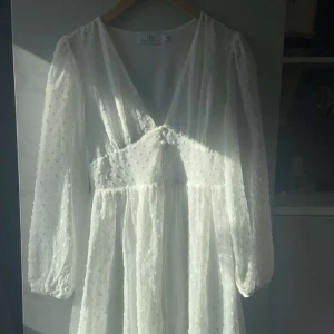 Vit kortklänning med prickigt mönster och volang - Superfin vit kortklänning med diskret prickigt mönster och volangdetaljer nertill. Djup v-ringning och långa ärmar med lätt puff. Perfekt för sommarens alla tillfällen!