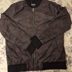 Svart camouflage bomberjacka från SMOG - Säljer en svart bomberjacka från SMOG med diskret camouflage-mönster. Jackan har dragkedja framtill, ribbade muddar och en ärmficka med dragkedja. Perfekt för dig som gillar streetwear och vill ha en stilren look.