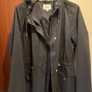 Mörkblå vindjacka med huva från Isolde - Säljer en mörkblå vindjacka från Isolde i storlek 36. Jackan har huva med randigt foder, dragsko i midjan och stora fickor framtill. Stängs med dragkedja och knappar. Perfekt för blåsiga dagar och enkel att matcha med olika outfits.