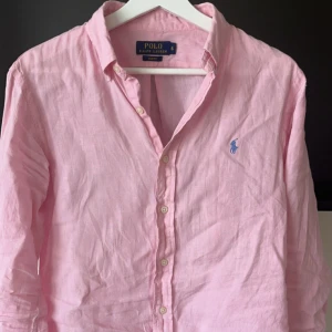 Rosa linneskjorta från Polo Ralph Lauren - Säljer en ljusrosa långärmad skjorta från Polo Ralph Lauren i linne. Skjortan har slim fit-passform, klassisk krage och knappar framtill. Broderad blå logga på bröstet. Perfekt för en fräsch och stilren look.
