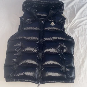 Svart dunväst från Moncler med huva - Snygg svart dunväst från Moncler med glansig finish och avtagbar huva. Västen har två dragkedjefickor framtill, quiltad design och klassisk Moncler-logga på bröstet. Perfekt för lager-på-lager och streetstyle. NFC-scan funkar. Använd en gång eftersom att den var för stor för mig. Passar perfekt till folk runt 170-175cm. Om ni har frågor är det bara att ställa!
