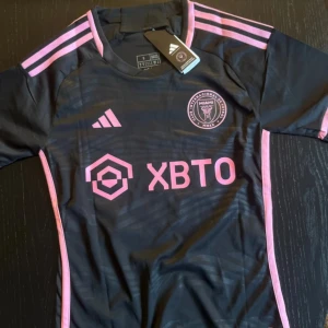 Inter Miami Messi #10 fotbollströja S - Inter Miami CF matchtröja  i svart med rosa detaljer och tryck. Messi #10 på ryggen, klubbmärke och sponsorlogga framtill. Klassisk rund hals, korta ärmar och tre rosa ränder på axlarna.