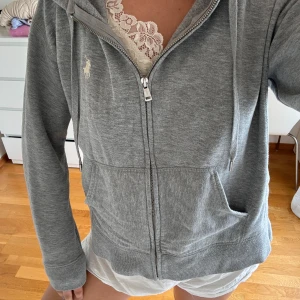 Grå hoodie från Polo Ralph Lauren - Grå hoodie med dragkedja från Polo Ralph Lauren. Klassisk modell med huva, snörning och kängurufickor framtill. Broderad logga på bröstet och ribbade muddar. Perfekt för chill dagar eller när du vill ha en clean och enkel look.