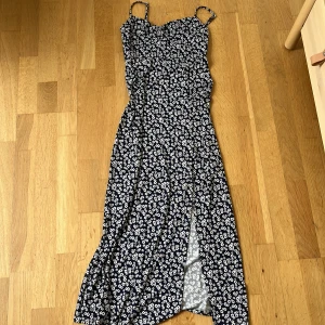 Blommig marinblå sommarklänning - Söt marinblå klänning med vita blommor och smala axelband. Klänningen har en slits framtill och figurnära passform upptill. Perfekt för varma dagar och har en lätt, flowy känsla.