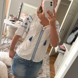 Vit blus med blåa detaljer - Superfin vit blus med blå broderade detaljer på ärmarna och framtill. Blusen har vida trekvartsärmar, knytning med blå tofsar vid halsen och avslappnad passform. Perfekt för sommaren eller för semester.