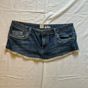 Jeanskjol med spetsdetaljer - Midjemått rakt över: 44 cm, längd: 23 cm Supersöt kort jeanskjol från Kut med ljus spetskant längst ner och broderade detaljer på fickorna. Klassisk blå denim med snygga kontrastsömmar och coola bakfickor med knappar. Perfekt för dig som vill ha en unik och trendig look.