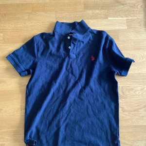 Marinblå pikétröja från U.S. Polo Assn. - Snygg marinblå pikétröja från U.S. Polo Assn. med klassisk krage, korta ärmar och knäppning framtill. Tröjan har röd broderad logga på bröstet och är tillverkad i mjuk bomull. Perfekt för en clean och sportig stil.