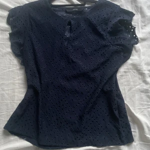 Mörkblå blus  - Supersöt mörkblå blus från Vero Moda med broderade hål i ett diskret mönster. Blusen har korta volangärmar och en liten öppning med knapp framtill. Perfekt för dig som gillar romantiska detaljer och en luftig känsla.