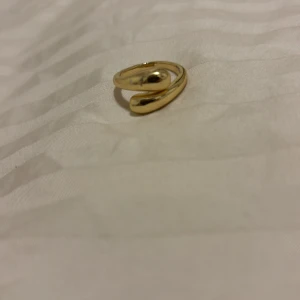Trendig guldig statementring - Säljer en snygg guldig ring med modern, öppen design och rundade droppformade ändar. Perfekt accessoar för att lyfta din outfit med en cool och stilren touch. Passar dig som gillar unika smycken med lite edge.