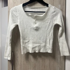 Vit ribbad långärmad tröja med knappar - Supersöt vit ribbad tröja med lång ärm och fyra knappar framtill. Tröjan är figurnära och har en lite croppad passform. Perfekt till jeans eller kjol för en clean och fräsch look.