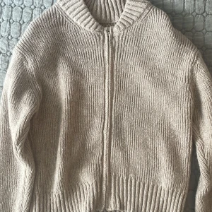 Beige stickad kofta med dragkedja - Mysig beige stickad kofta med grov ribbad krage och dragkedja framtill. Långärmad modell med breda muddar vid ärmslut och nederkant. Perfekt för lager-på-lager och chill dagar.