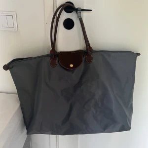 Grå shoppingväska från Longchamp - Stor och rymlig shoppingväska från Longchamp i grått med bruna skinnhandtag och lock. Väskan har guldfärgade detaljer och dragkedja upptill. Perfekt för dig som vill ha plats för allt du behöver under dagen. Dragkedjan är trasig.