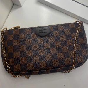 Louis Vuitton Brun väska  - Säljer en ikonisk axelväska från Louis Vuitton i klassiskt brunt och mörkbrunt rutigt mönster. Väskan har guldfärgad kedja och dragkedja upptill. Perfekt storlek för det viktigaste och riktigt snygg att bära på axeln! Bra skick då den inte har används många gånger. 