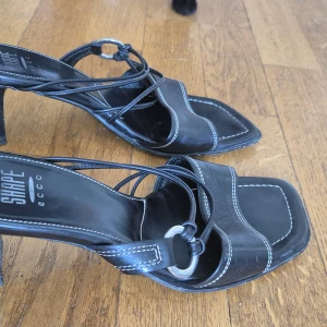 Svarta mules från Ecco - Svarta mules sandaler med vita konstrastsömmar från Ecco storlek 38 och 5cm klackar. Skorna har smala remmar och en metallring som detalj. Tillverkade i skinn och i fint skick. Ecco är kända för att göra väldigt bekväna skor, vilket dessa även är! Tyvärr lite för små för mig. Passar en normal 38, jag lutar mer åt 39.