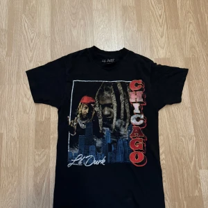Svart Lil Durk Chicago t-shirt - Svart t-shirt med tryck av Lil Durk och texten 'CHICAGO' i stora röda bokstäver längs sidan. Stadssilhuett i blått och vitt nertill. Klassisk passform och rund hals. Perfekt för dig som gillar streetwear och hiphop vibes/Vintage stil