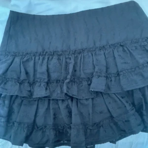 Svart volangkjol från H&M Divided - Supersöt svart kortkjol från H&M Divided med flera lager volanger och diskret broderat mönster i tyget. Kjolen har en lätt struktur och ger en cool, lekfull vibe. Perfekt att styla med en croppad topp eller oversized hoodie. 74cm i midjemått helt ny aldrig använd!!!!