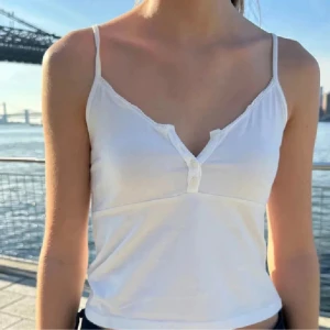 Vit linnetopp från Brandy Melville - Supersöt vit linnetopp från Brandy Melville med smala axelband och v-ringning. Toppen har tre små knappar framtill och en diskret spetskant längs urringningen. Perfekt för varma dagar och enkel att styla till jeans eller kjol.