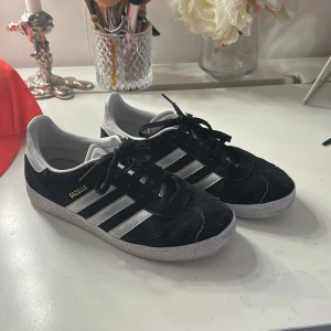 Adidas Gazelle svarta sneakers mocka - Svarta Adidas Gazelle sneakers i mocka med klassiska vita ränder och vit sula. Skorna har snörning och rund tå, samt diskret guldfärgad Gazelle-text på sidan. Perfekta för en clean och sportig look.