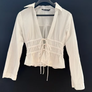 Vit snörad skjorta från Zara - Vit skjorttopp från Zara med lång ärm, djup v-ringning och snygga snörningsdetaljer framtill. Toppen har markerad midja med rynk och lätt croppad passform. Perfekt för dig som gillar trendiga och feminina plagg.