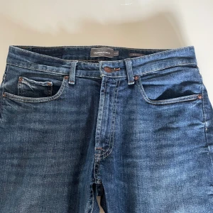 Blå jeans från Dressmann regular fit - Klassiska blå jeans från Dressmann i regular fit med fem fickor och snygga kontrastsömmar. Jeansen har en mörkblå tvätt och brunt läderpatch bak i midjan. Perfekta för dig som gillar en tidlös och enkel stil.