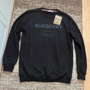 Svart Burberry sweatshirt med tryck - Svart sweatshirt från Burberry med stort tryck på bröstet och klassisk rund hals. Tröjan har långa ärmar, ribbade muddar och är tillverkad i mjuk bomull. Perfekt för en stilren och trendig look med exklusiv känsla.