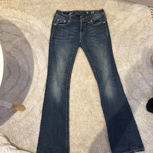 Miss Me jeans med broderi och nitar - Snygga blå jeans från Miss Me med bootcut-modell. Baksidan har detaljerade broderier, nitar och strass på fickorna samt ett stort M-emblem. Jeansen har låg midja och coola slitningar för en edgy look. Perfekta för dig som gillar statement-jeans ger dig en känsla av 2000s stil 