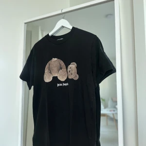 Svart Palm Angels t-shirt med björntryck - Svart t-shirt från Palm Angels med ikoniskt björntryck framtill och stor vit logga på ryggen. Klassisk passform, rund hals och korta ärmar. Tillverkad i mjuk bomull, perfekt för en streetwear-look.