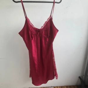 Röd satin natt klänning med spets - Snygg röd kort natt klänning i satin med tunna axelband som går att justera och v-ringning. Klänningen har spetsdetaljer vid bysten och längs sidorna samt en liten rosett framtill. Orginalpriset va ca 460kr 