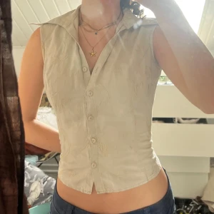 Beige ärmlös skjorta med broderi - Snygg beige skjorta utan ärmar med broderade detaljer och klassisk krage. Skjortan har figurnära passform och knäpps framtill med knappar. Perfekt för dig som vill ha en stilren men ändå unik look.