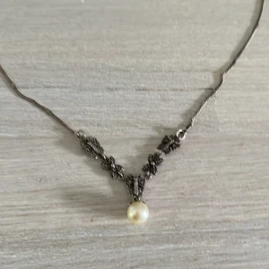 Silverhalsband Alton med pärla - Unikt halsband i äkta silver (925) från Albrekts guld, med detaljerat blommönster och en hängande vit pärla. Kedjan är tunn och smidig, och smycket levereras i original ask. Perfekt för dig som gillar klassisk och elegant stil.