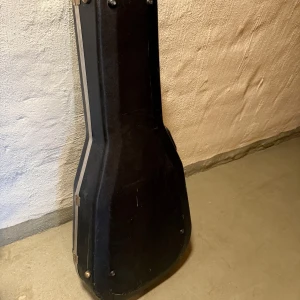 Svart hårt gitarrfodral - Säljer ett svart hårt gitarrfodral med plyschfoder på insidan. Fodralet har metallspännen och handtag på sidan. Formen är anpassad för akustisk gitarr och insidan är mjuk och skyddande. Perfekt för att transportera eller förvara din gitarr.