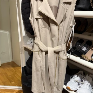 Beige ärmlös klänning Calvin Klein - Snygg beige ärmlös kortklänning från Calvin Klein med markerad krage, dragkedja framtill, fickor med gulddetaljer och matchande bälte i midjan. Perfekt för dig som gillar stilrena och moderna plagg med coola detaljer.