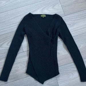 Svart omlottopp från Vila - Säljer en svart ribbad omlottopp från Vila i storlek M (passar dock mig som har strl S). Toppen har lång ärm, djup v-ringning och asymmetrisk nederkant. Den är figurnära och tillverkad i ett stretchigt material som sitter snyggt på kroppen.