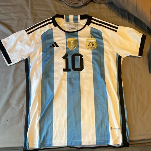 Argentina Messi 10 fotbollströja Adidas M - Säljer en officiell Argentina fotbollströja från Adidas i storlek M, med Messi och nummer 10 på ryggen. Tröjan är randig i vitt och ljusblått, har korta ärmar och broderat AFA-märke med tre stjärnor. Tillverkad i lätt och ventilerande polyester (AEROREADY).