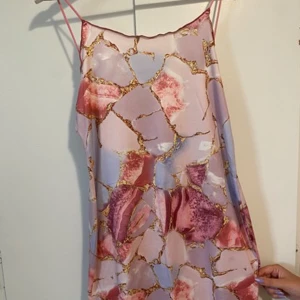 Pastellfärgad omlottklänning med mönster - Säljer en superfin kortklänning med tunna axelband och ett unikt mönster i rosa, lila och guld. Klänningen har en omlottkänsla och är tillverkad i ett glansigt, lätt material som känns lyxigt. Använt en gång