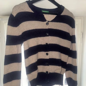 Randig kofta från United Colors - Beige och mörkblå randig kofta från United Colors of Benetton. Klassisk modell med rund hals, långa ärmar och svarta knappar framtill. Mjuk stickad känsla som passar perfekt till jeans eller kjol.