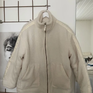Beige teddyjacka med dragkedja S - Mysig beige teddyjacka med hög krage och dragkedja framtill. Jackan har två stora fickor och en quiltad insida. Snygga detaljer med ljusa band längs axlarna. 
