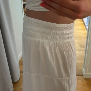 Vit långkjol med smock i midjan - Superfin vit långkjol med bred smock i midjan som ger en skön och flexibel passform. Kjolen är luftig och har ett lätt, flowy tyg som passar perfekt till varma dagar. Enkel och stilren design som är lätt att matcha med olika toppar. Köpt på ginatricot och är stretchig så passar även dig som har lite större storlek än 146/152, i längden är den lite för kort på mig som är 164cm ❤️skriv vid funderingar