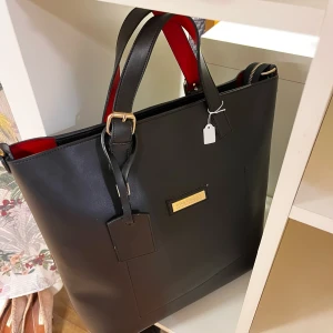 Svart axelväska från Burberry - Stilren svart axelväska från Burberry med guldiga metalldetaljer och röd insida. Väskan har dubbla handtag, justerbart spänne och en klassisk Burberry-logga framtill. Tillverkad i slätt skinn. Köpt på secondhand så oklart om den är äkta därav priset! 