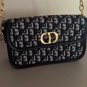 Dior axelväska i blått monogram - Säljer en snygg axelväska från Dior med klassiskt blått och vitt monogrammönster. Väskan har guldfärgat spänne med Dior-logga och kedjerem i metall. Materialet är en mix av textil och skinn, perfekt för dig som vill ha en ikonisk accessoar med lyxig känsla.