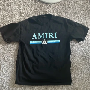 Amiri t-shirt -  Oanvänd Svart t-shirt  från AMIRI i bomull med stort blått och vitt logotryck på bröstet. Klassisk passform med rund hals och korta ärmar. Kan även sänka vid snabb affär. 
