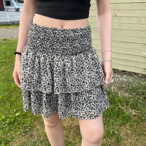 Volangkjol - Supersöt kort kjol med leopardmönster i svart och beige. Kjolen har bred smock i midjan för en bekväm passform och dubbla volanger som ger en flowy och trendig look. Perfekt för dig som vill sticka ut lite extra i sommar.