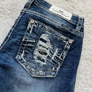 Miss Me blå bootcut jeans med detaljer - Snygga blå jeans från Miss Me med bootcut passform. Jeansen har slitna detaljer och dekorerade bakfickor med nitar, broderier och glittriga stenar. Tydliga kontrastsömmar och coola knappar ger extra edge. Perfekta för dig som gillar statement-jeans.