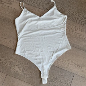 Vit bodysuit från H&M med smala axelband - Säljer en stilren vit bodysuit från H&M i mjuk bomullsmix. Modellen har smala axelband, v-ringning och knäppning i grenen. Perfekt att styla med jeans eller kjol för en clean look.
