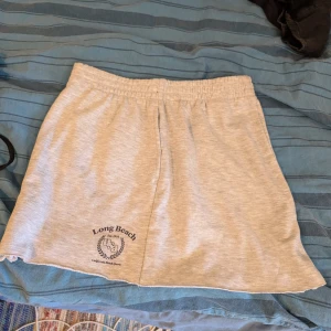 Grå kortkjol från H&M Divided - Sportig ljusgrå kortkjol från H&M Divided med tryck 'Long Beach USA' framtill. Kjolen har resår i midjan för skön passform och är gjord i mjuk bomullsblandning. Perfekt för chill dagar eller strandhäng.