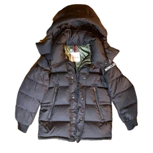 Sällsyn Moncler dunjacka med reflexdetalj - Säljer en svart Moncler dunjacka med avtagbar huva och stor reflekterande M-logga på ryggen. Jackan har dragkedjor på fickorna, ribbade muddar och glansig insida i olivgrönt. Perfekt för kalla dagar och riktigt snygg streetstil. Storlek 2 